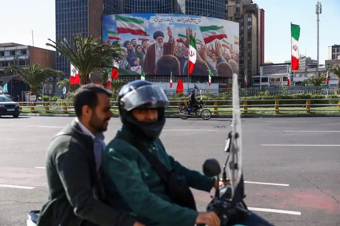 People ride on a motorbike past a billboard featuring an image of the late Supreme Leader of Iran, Ayatollah Ali Khamenei.