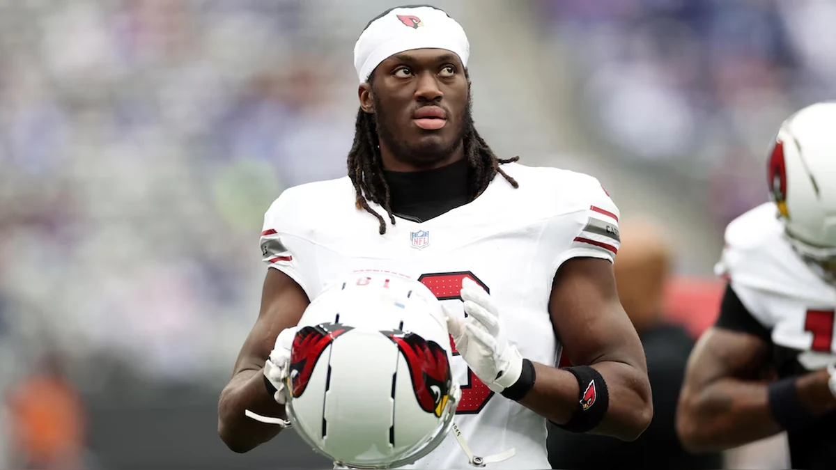 Arizona Cardinals player in a white uniform and white headband holding his helmet on the football field.